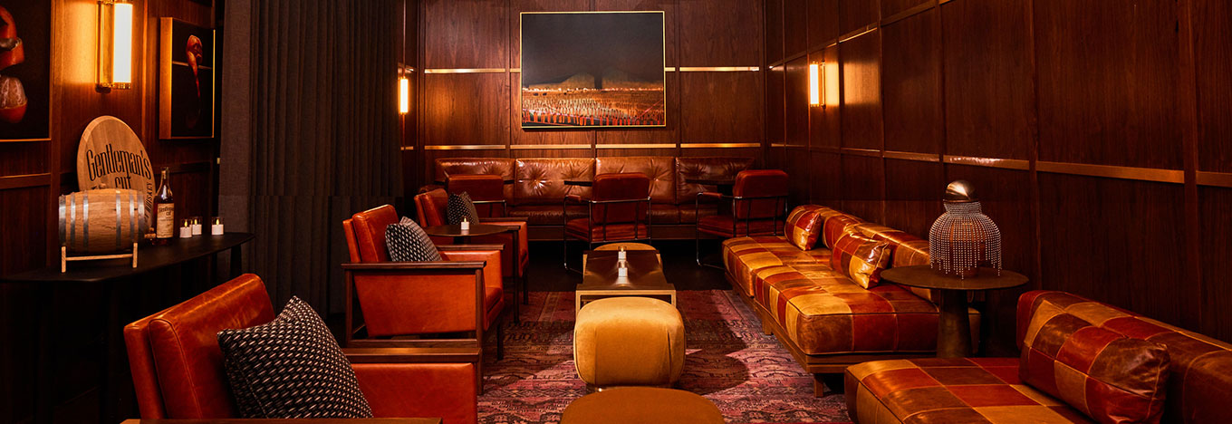 A cozy, dimly lit lounge with brown leather sofas, wooden paneled walls, and a patterned rug. A painting hangs on the back wall above leather chairs. Accent lighting and throw pillows add a warm, inviting atmosphere.