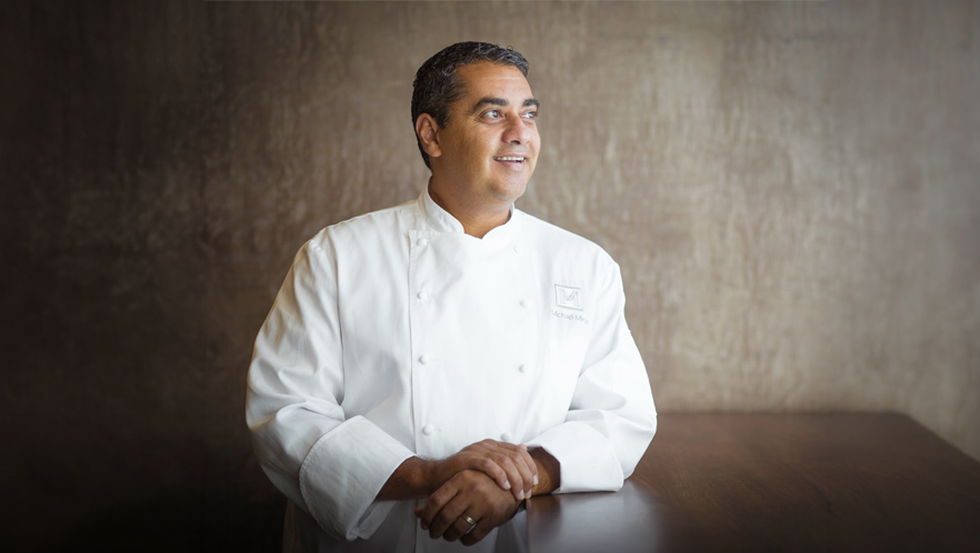 Chef Michael Mina in a white double-breasted coat stands behind a wooden table, smiling and looking to the right. The background is a muted, textured brown wall, and the lighting softly highlights his face and uniform.