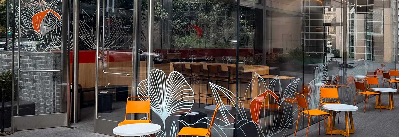 A modern café with glass walls features orange chairs and small round white tables outside. Large white line art flower designs decorate the glass. Inside, wooden tables, high chairs, and wine glasses are visible. Greenery is reflected in the glass.