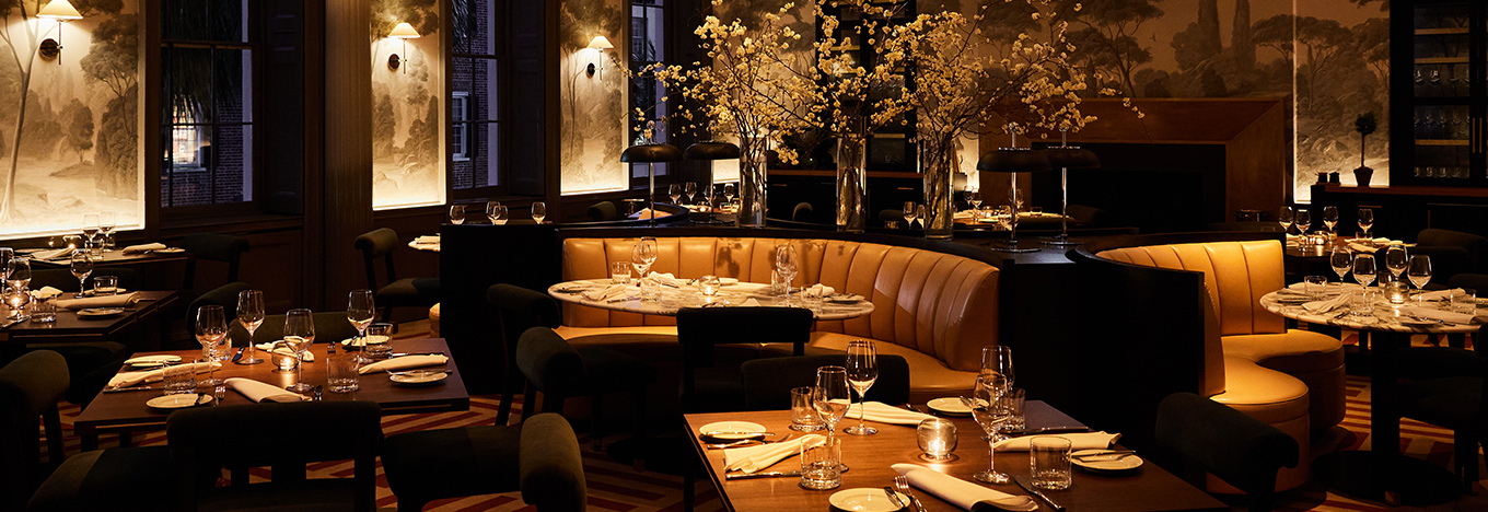 Elegant restaurant interior with dim, warm lighting. Tables are set with white napkins, glasses, and plates. Curved yellow booth seating surrounds round tables with centerpiece vases of yellow flowers. Large windows and mural walls add sophistication.