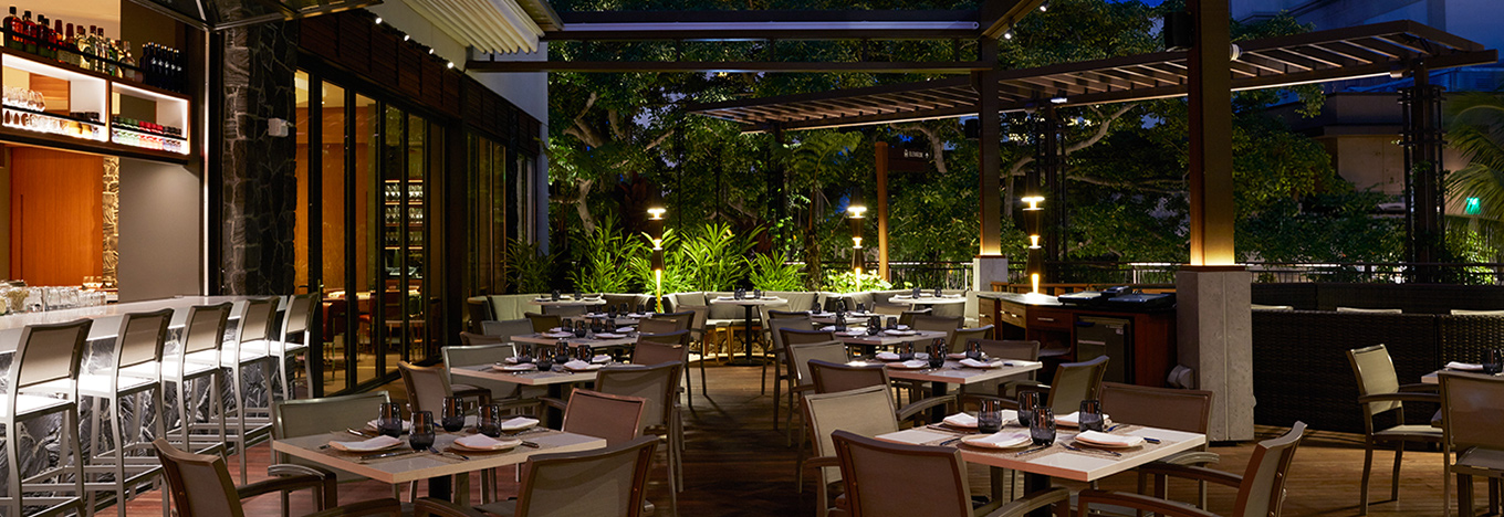Modern outdoor restaurant at night with wooden flooring, tables set with plates and glasses, beige chairs, bar area with stools on the left, and green plants and trees providing a lush backdrop under warm ambient lighting.
