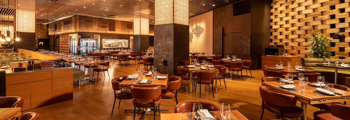 Upscale restaurant with warm lighting, brown leather chairs, and neatly set tables with glasses and napkins. Wooden floors and walls, modern decor, vertical light panels, and a mix of booth and chair seating create an inviting atmosphere. No people are present.
