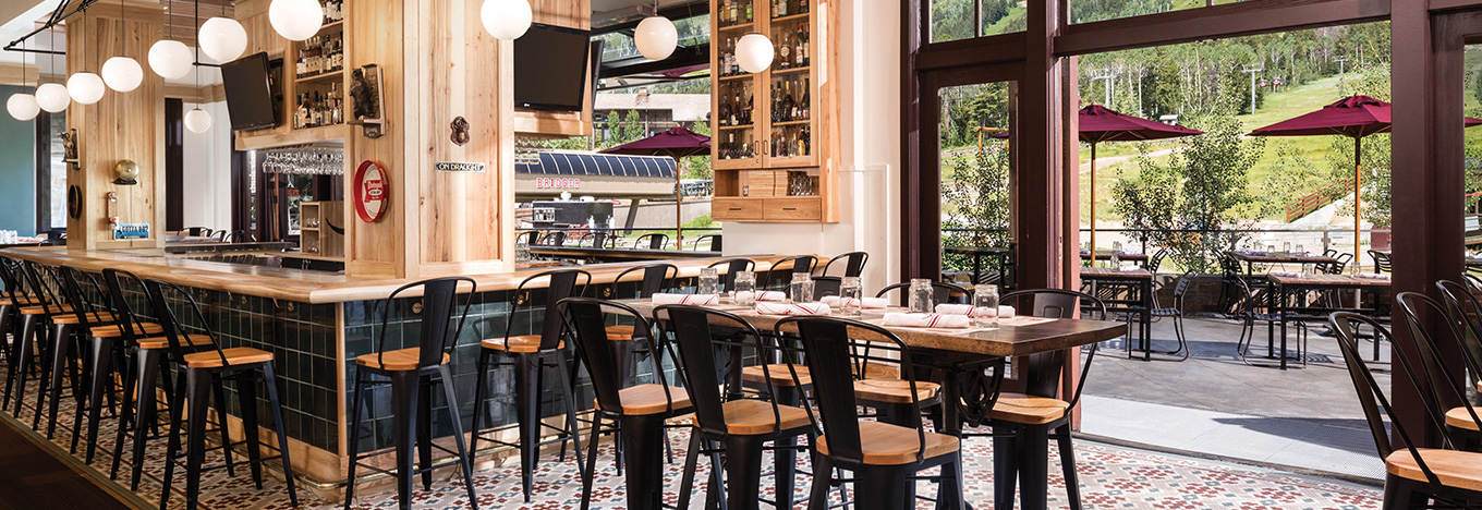 Modern restaurant interior with a long bar, high black chairs with wooden seats, tables set for dining, and large windows opening to a patio with red umbrellas and a view of green hills and trees. The space is bright, airy, and inviting.