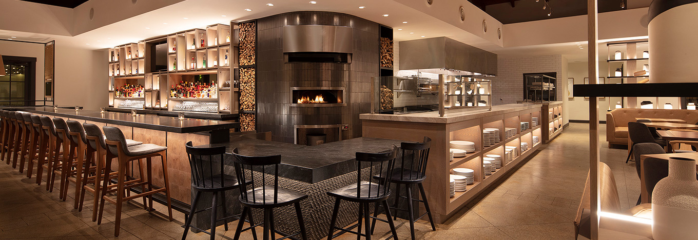 Modern restaurant interior with a large open kitchen and wood-fired oven, surrounded by countertops and shelves displaying plates and decor. Barstools line the counters, and cozy booths and tables fill the softly lit, spacious dining area.