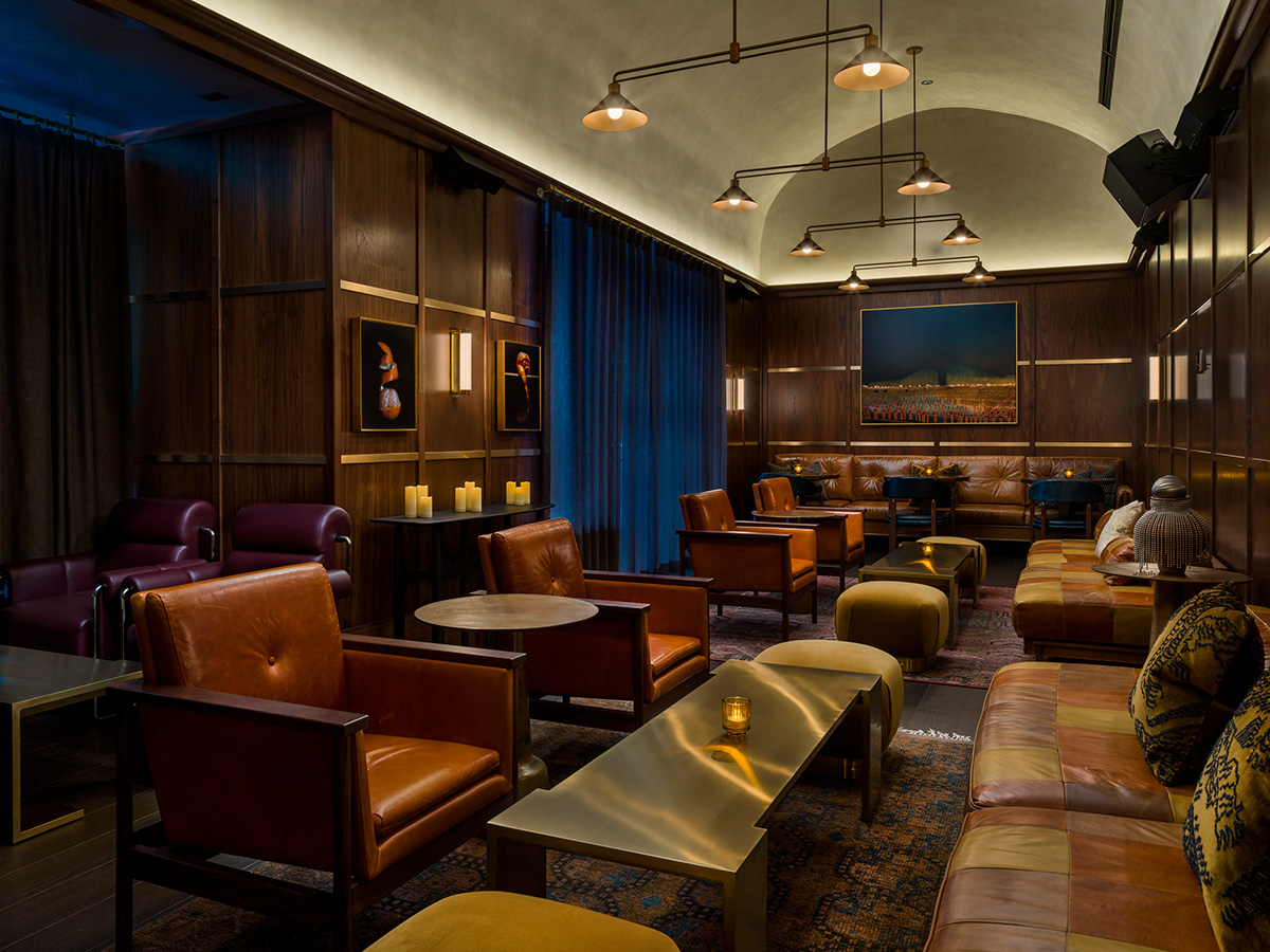 A dimly lit lounge with brown leather chairs, sofas, and wooden walls. Warm lighting hangs from the ceiling. Artworks and candles decorate the space. Blue curtains cover tall windows. A large painting is mounted on the far wall above a brown couch.