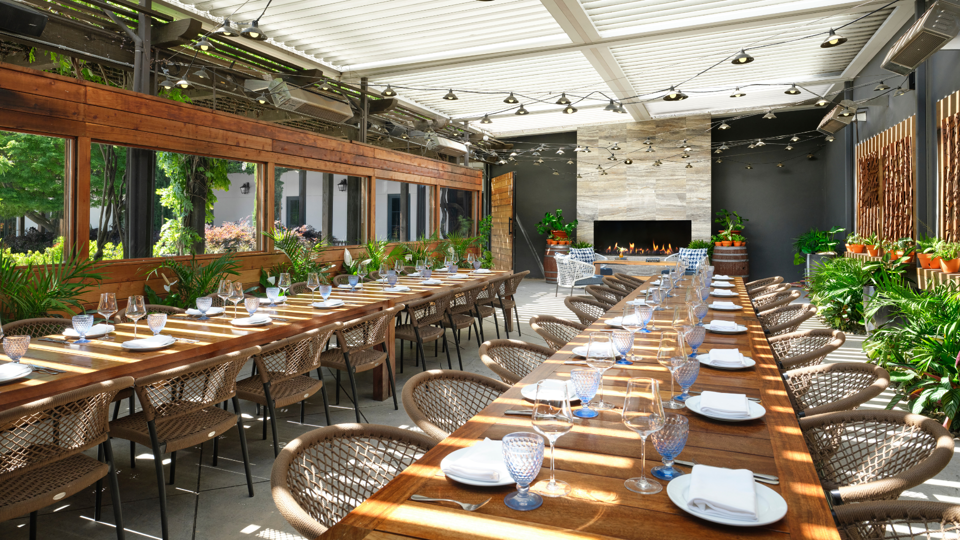 A bright, modern dining area with two long wooden tables set with plates, glasses, and napkins. Wicker chairs surround the tables. Green plants decorate the space, and a stone fireplace sits at the end under a glass roof with exposed beams.