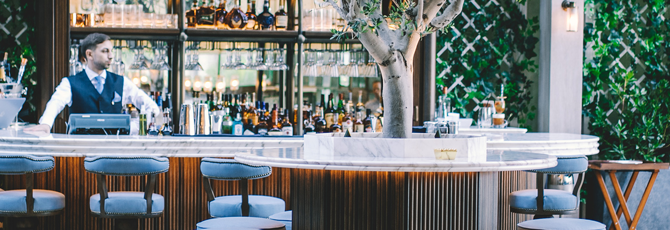 A bartender in a white shirt and vest stands behind a modern marble bar, surrounded by bottles, glassware, and a central tree. Plush blue barstools line the counter, and lush green plants create a stylish, upscale outdoor atmosphere.