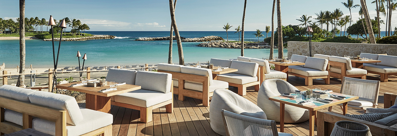 An outdoor restaurant patio with wooden tables and cushioned seating overlooks a calm, turquoise bay lined with palm trees. Tiki torches are scattered around, and the sky is bright and clear, creating a relaxed, tropical atmosphere.