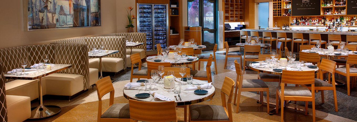 Elegant restaurant interior with round and square tables set with glasses, plates, and napkins. Wooden chairs surround tables; cushioned booth seating lines the left wall. A well-lit bar with bottles and stools is on the right. Art decorates the beige walls.