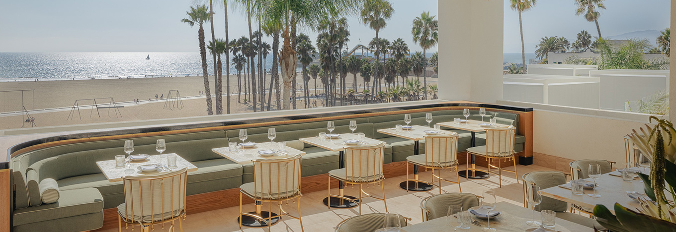 An elegant beachfront restaurant with pastel seating, gold-accented chairs, and set tables overlooks a sandy beach lined with palm trees. The ocean and a sailboat are visible in the background under clear skies.