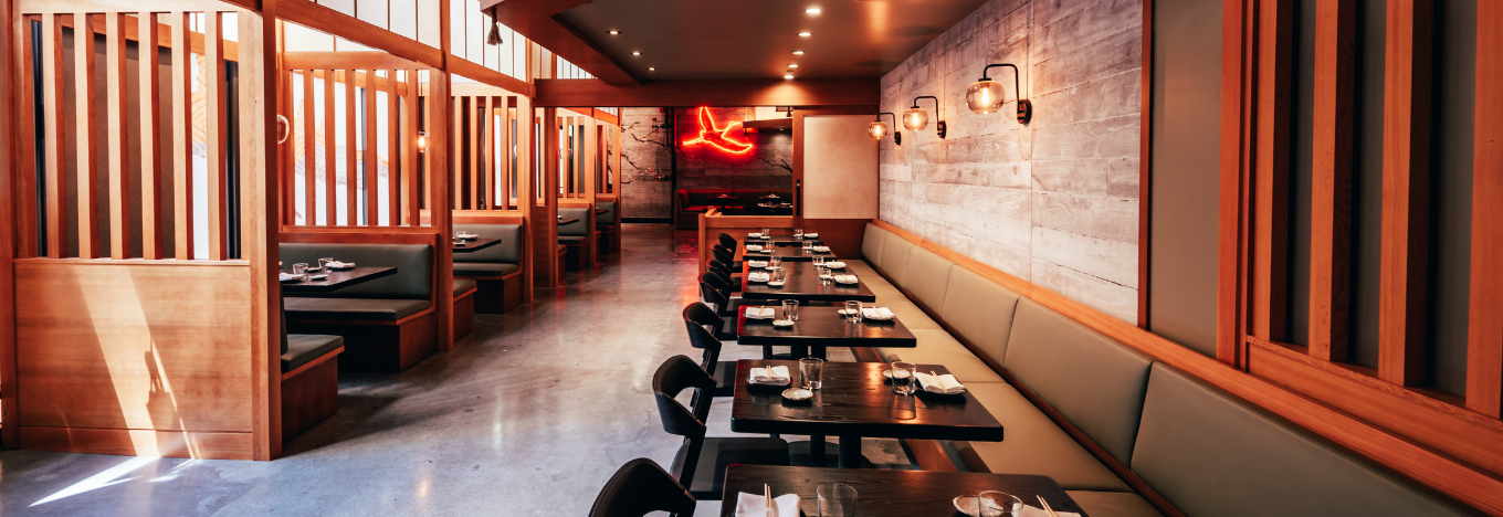 Modern restaurant interior with wooden partitions, polished concrete floor, and soft lighting. Black tables and chairs are arranged along a long olive-green bench on the right, with wall-mounted lights and a glowing red neon bird sign in the background.