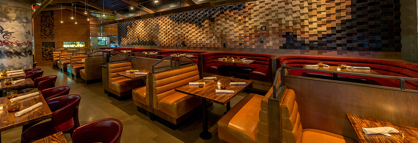 A modern restaurant interior with amber leather booths, wooden tables set with napkins, dim lighting, and a textured geometric wood panel wall. The atmosphere is warm and inviting, with artistic wall details and a cozy, upscale vibe.