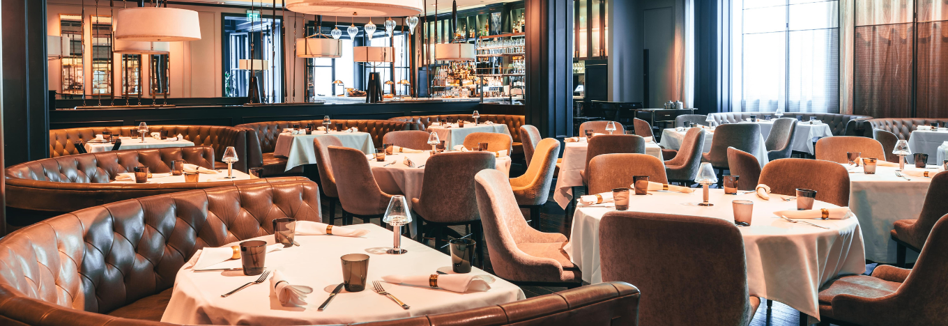 A stylish, upscale restaurant interior with round tables covered in white cloths, brown leather booths, beige upholstered chairs, and elegant table settings. Large mirrors, modern hanging lights, and a well-stocked bar are visible in the background.