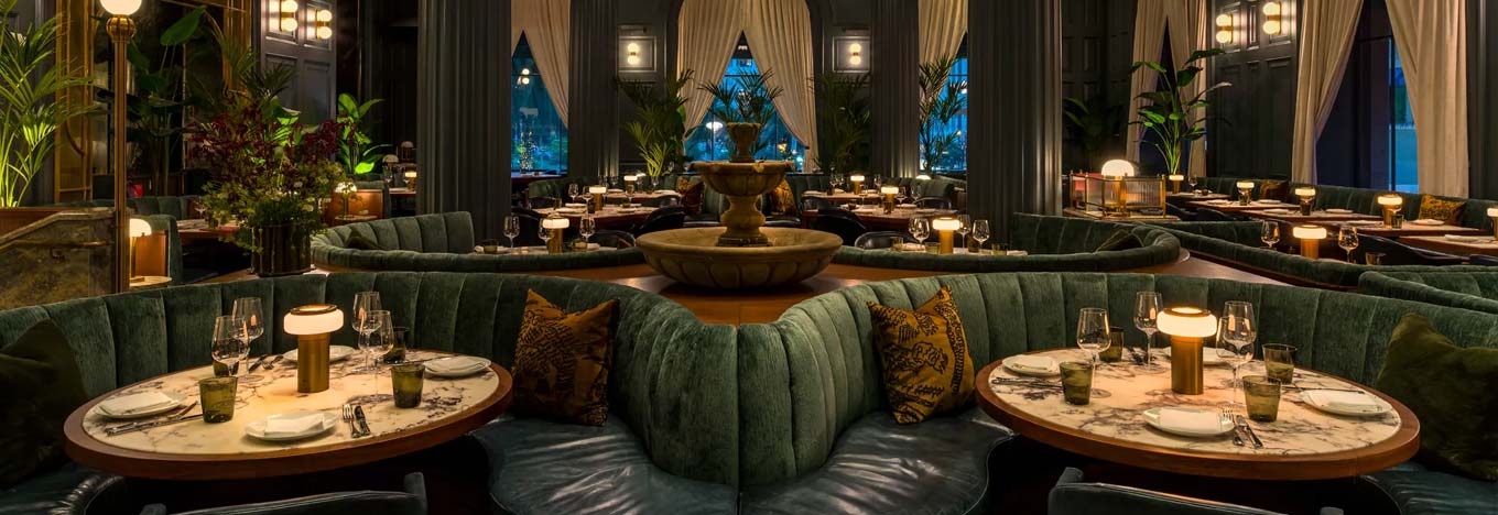 Elegant restaurant interior with round marble tables, green velvet curved booths, dim table lamps, and a central stone fountain. Large windows with sheer drapes, lush plants, and soft, ambient lighting create a warm and inviting atmosphere.
