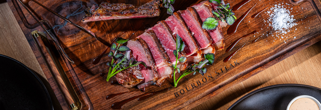 Sliced medium-rare steak is arranged on a dark wooden cutting board branded with BOURBON STEAK. The meat is garnished with sprigs of microgreens and a pinch of coarse sea salt, with streaks of dark sauce drizzled across the board.