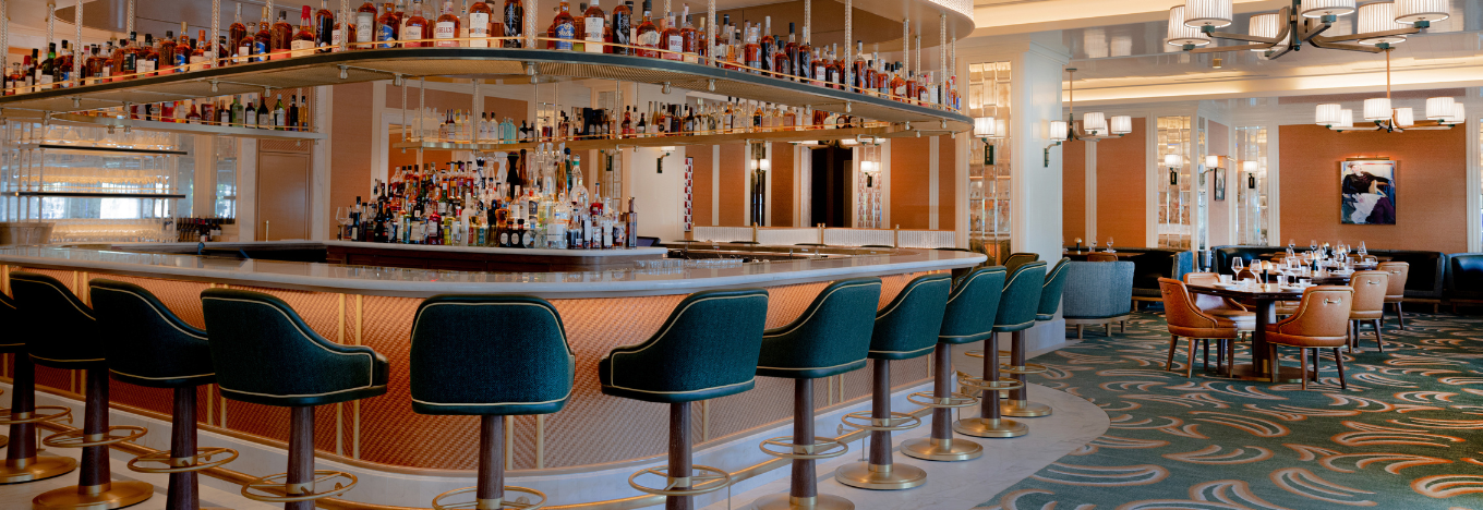 Chic restaurant interior with a central bar lined by green upholstered barstools. The bar displays shelves of liquor bottles above and behind it. Elegant dining tables with chairs are set nearby. Soft lighting and art deco touches create a modern, inviting atmosphere.