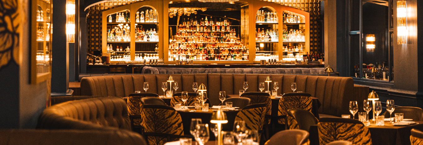 A warmly lit upscale restaurant with curved booths, zebra-patterned chairs, set tables with glassware, and a well-stocked bar in the background featuring shelves of bottles and soft golden lighting. The ambiance is elegant and inviting.