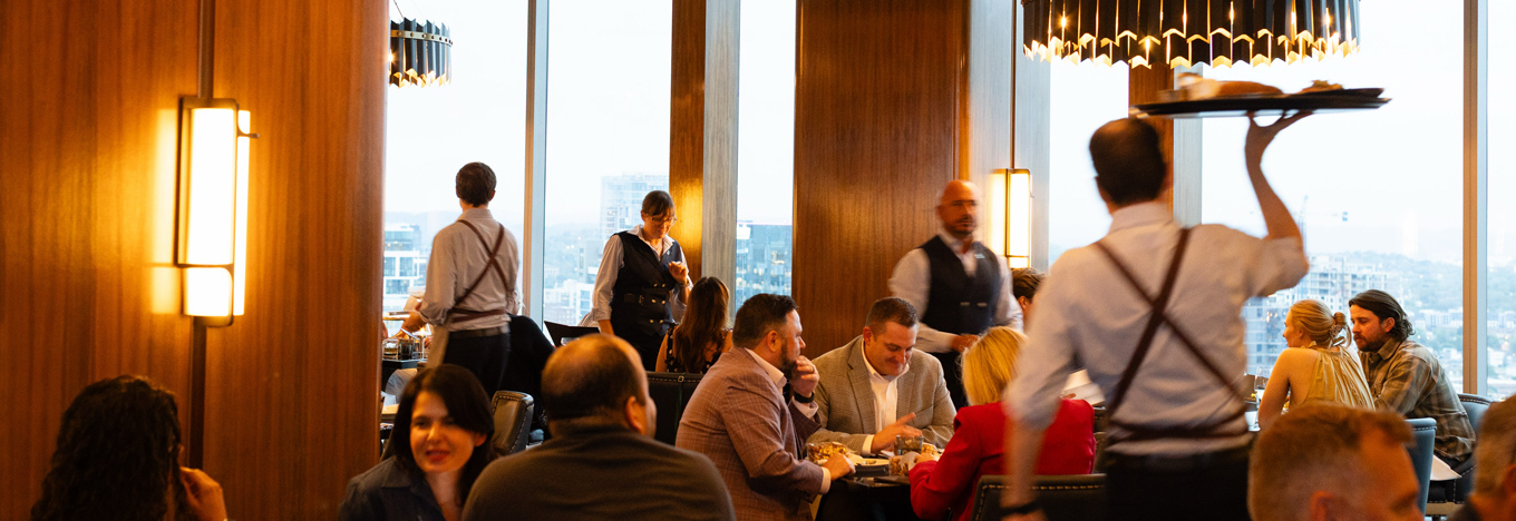 A server carries a tray above their head in a busy, high-rise restaurant with large windows. Diners sit at tables in the foreground, while other staff in uniform attend to the room.