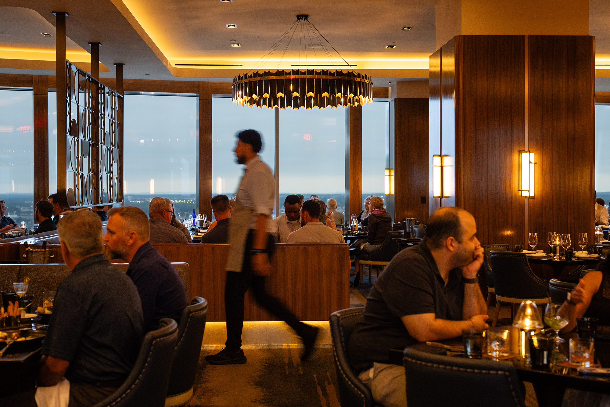 A server walks through a dimly lit, upscale restaurant with floor-to-ceiling windows overlooking a city at dusk. Patrons are seated at tables throughout the dining room, some in booths and others at individual tables.