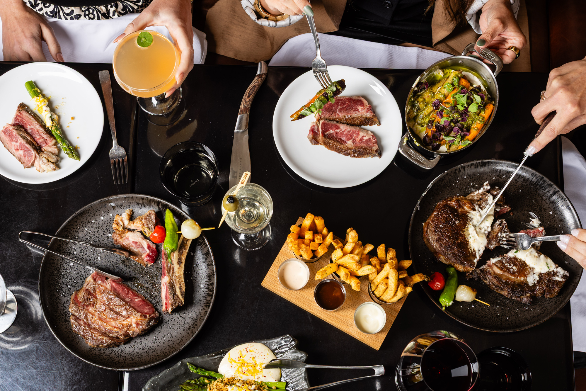 A top-down view of a dinner table featuring several plates of steak, roasted vegetables, and fries. Hands are visible using forks and knives to cut into the meat and serve from shared dishes, accompanied by glasses of cocktails and wine.