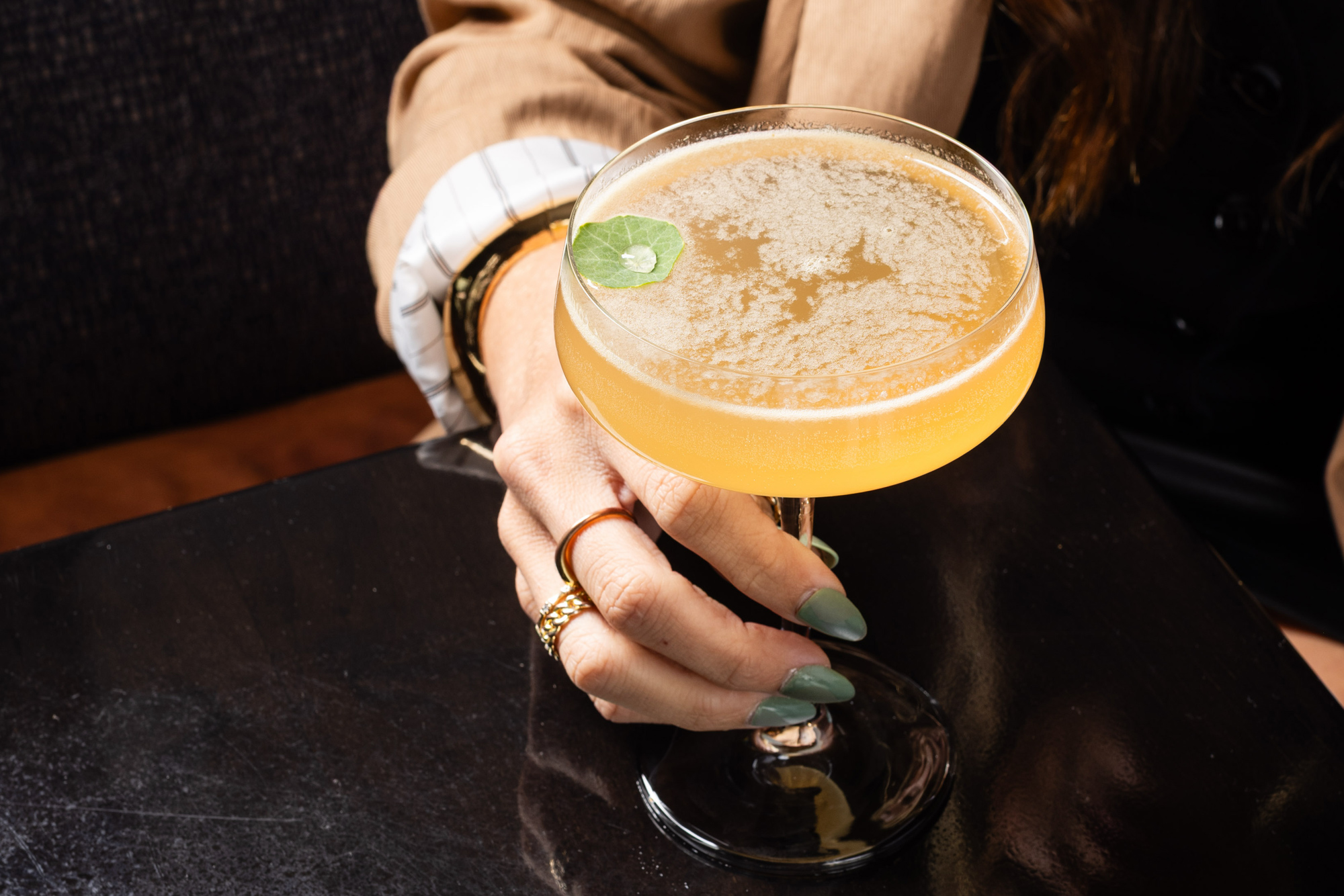 A hand holds a yellow cocktail in a coupe glass, garnished with a single small green leaf. The person is wearing several gold rings and green nail polish.