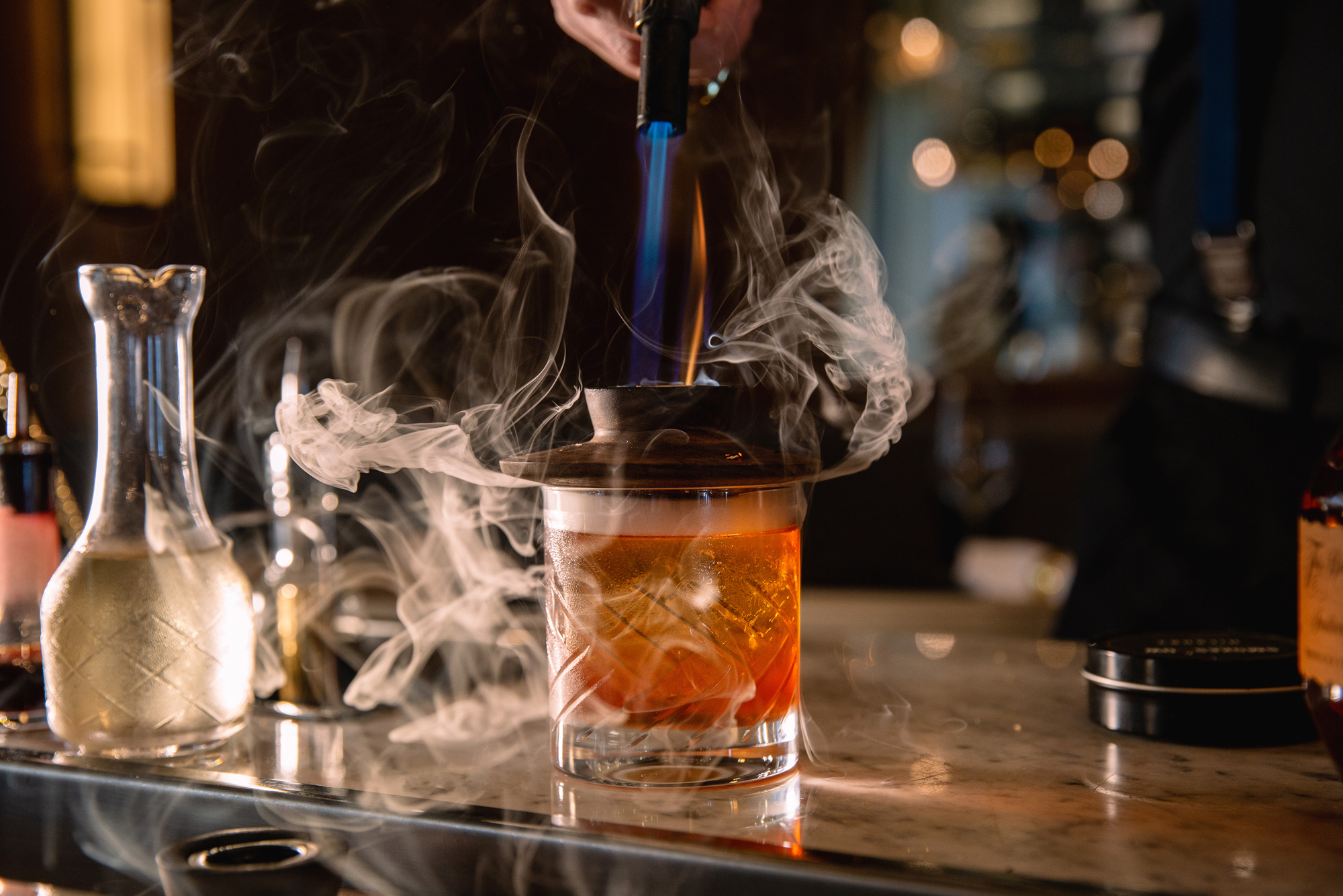 A bartender uses a torch to smoke a cocktail in a short, patterned glass. Swirls of smoke rise from the drink as it sits on a marble bar top.