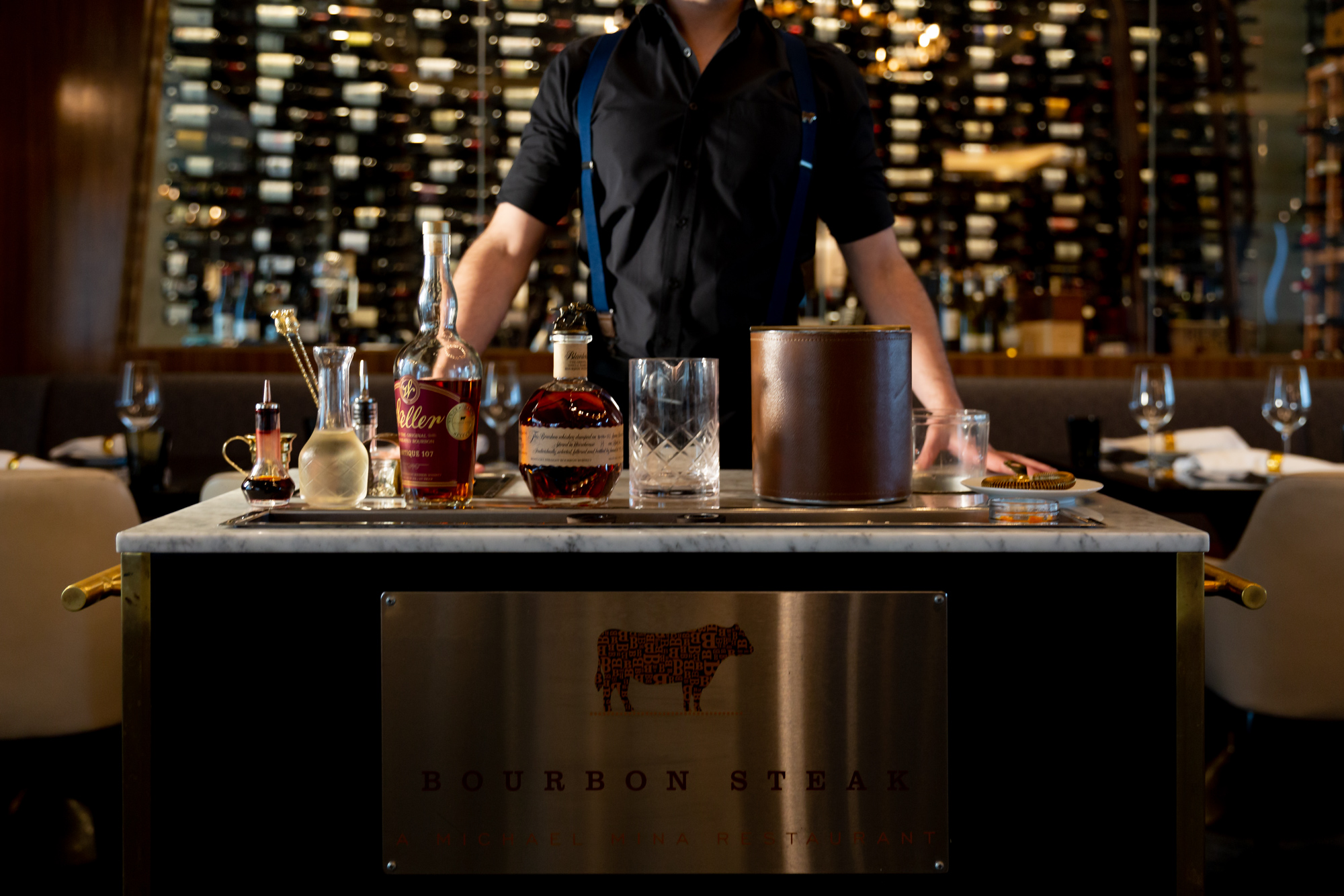 A bartender stands behind a mobile bar cart featuring bottles of Weller and Woodford Reserve bourbon. The cart features a metal plaque with a cow silhouette and the text Bourbon Steak a Michael Mina Restaurant in front of a wine cellar.