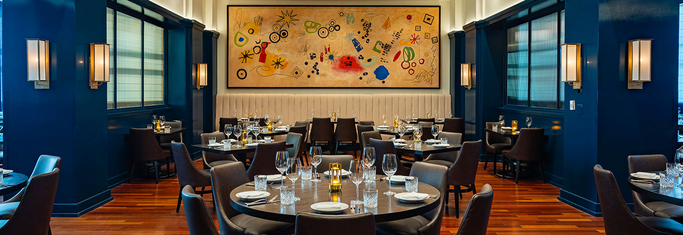 Elegant restaurant interior with rich blue walls, wooden floors, and round tables set with glassware and plates. Modern artwork with abstract shapes and bright colors hangs above tan banquette seating, creating a sophisticated yet inviting atmosphere.