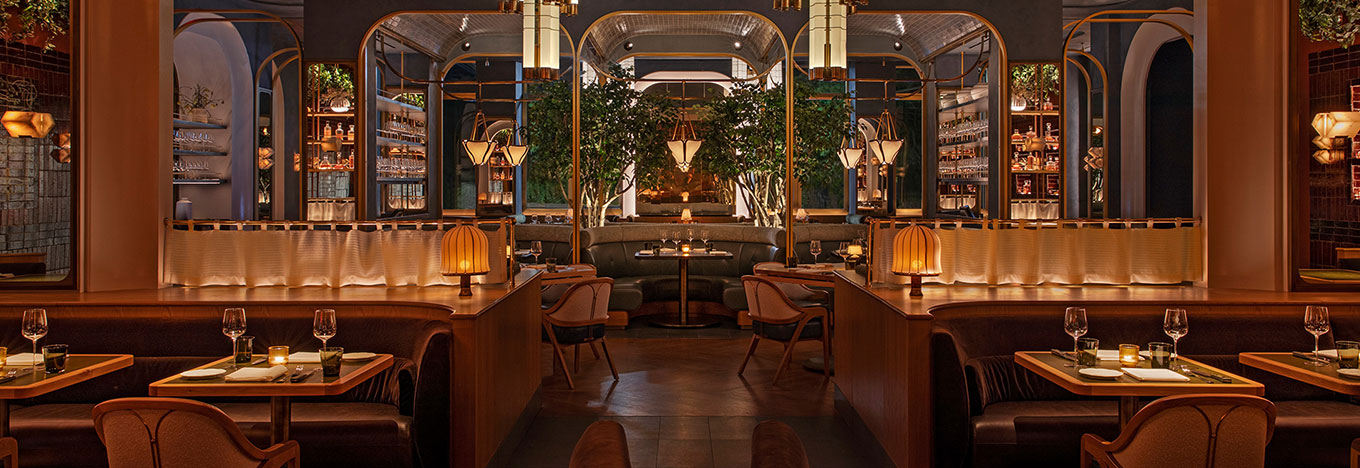 Elegant restaurant interior with warm lighting, round tables set with glasses and plates, cushioned booths and chairs, trees in the center, shelves with bottles and glassware, and soft amber lamps creating a cozy, upscale ambiance.