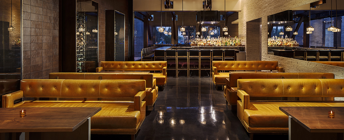 A modern, dimly lit bar interior with yellow-brown leather booths, wooden tables, and a glossy black floor. A mirrored wall reflects the bar, which is lined with high stools and shelves stocked with bottles. Small candles sit on some tables.