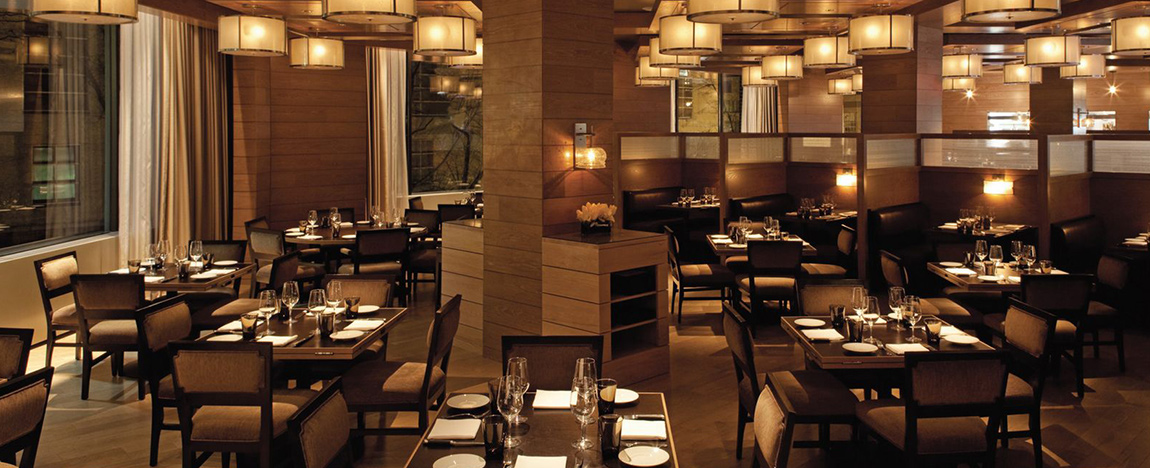 A warmly lit upscale restaurant with wooden walls and columns, neatly set dark tables, and cushioned chairs. Modern pendant lights hang from the ceiling, and large windows line one wall. The room is empty, with place settings ready for guests.