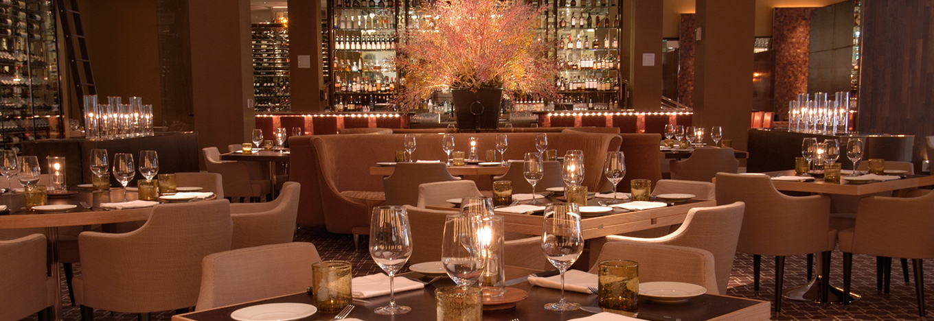 Elegant restaurant interior with beige chairs, wooden tables set with wine glasses, plates, and candles. Large floral arrangement and backlit shelves with bottles behind a central curved booth. Warm, ambient lighting creates a cozy atmosphere.