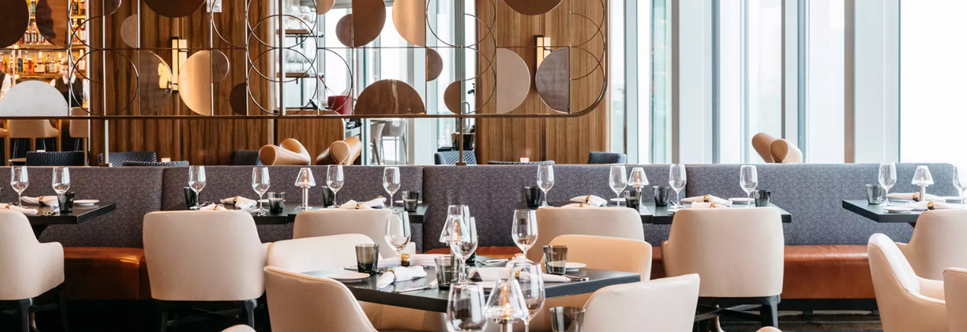 A modern, upscale restaurant with cream chairs, dark tables set with glasses and napkins, and a padded bench along the wall. Geometric screen dividers and large windows let in natural light. The atmosphere is elegant and inviting.