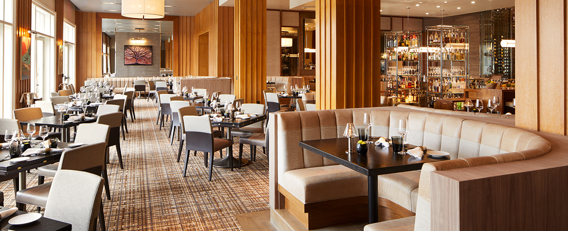 A modern restaurant interior with tan upholstered booths, dark tables, and beige chairs. Wooden columns and patterned carpet add warmth. Place settings with glasses and napkins are on tables. Floor-to-ceiling windows and a stocked bar are in the background.