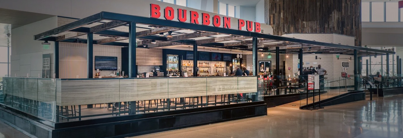 A modern bar called Bourbon Pub with bold red letters on the sign. The open layout shows a counter with bottles and seating inside, glass barriers around the perimeter, and a few patrons inside. The area is well-lit with a polished floor.