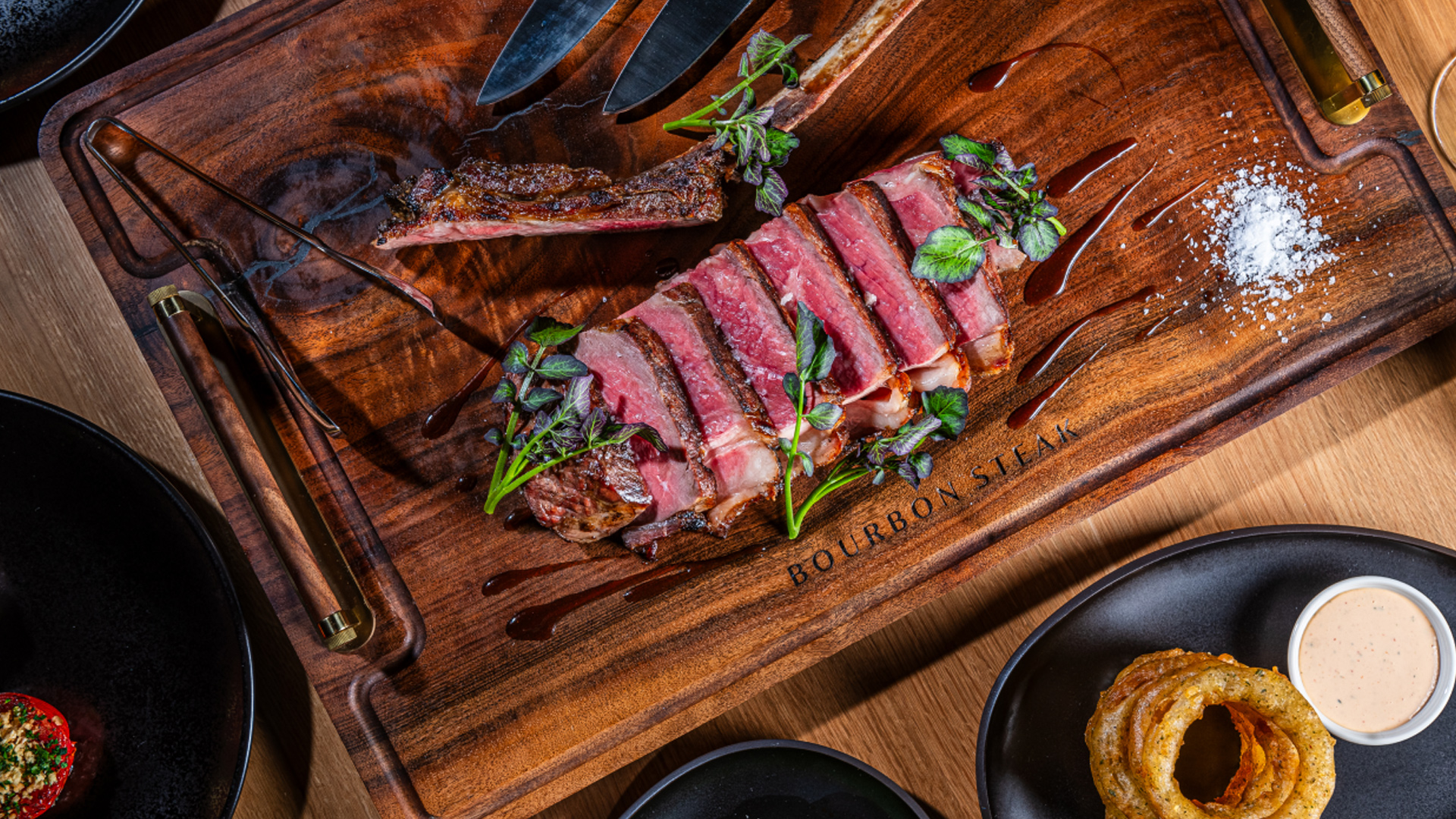 A sliced medium-rare steak is served on a wooden board labeled Bourbon Steak, garnished with herbs. Salt and sauce are on the side, along with onion rings on a black plate and a knife and fork resting nearby.