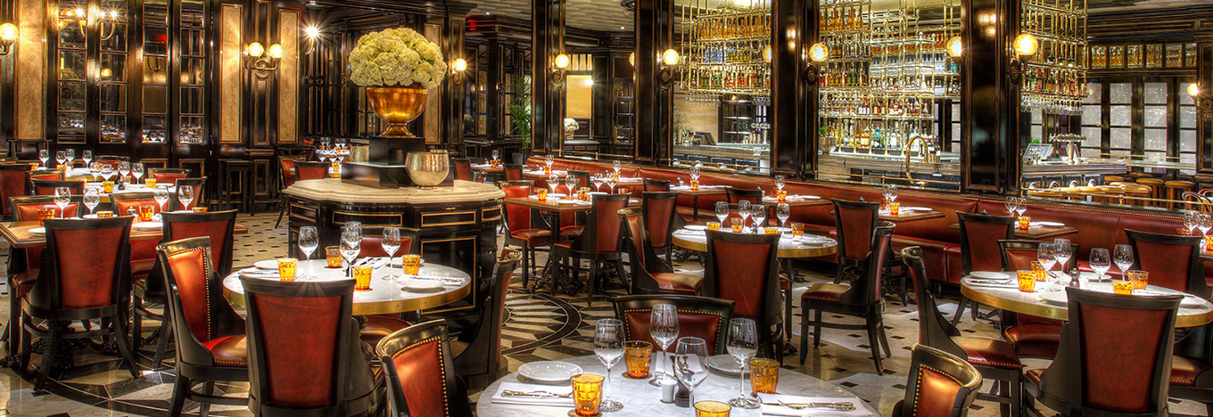 Elegant restaurant interior with round marble tables, red leather chairs, and amber glassware. A large floral arrangement sits in the center. Dark wood, mirrored walls, and shelves of bottles create a luxurious atmosphere with warm lighting.