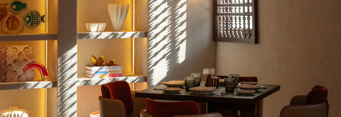A cozy dining area with a square table set for four, surrounded by cushioned chairs. Sunlight casts patterned shadows on the wall through a wooden lattice, while shelves display colorful ceramics, art pieces, and books, all warmly lit.