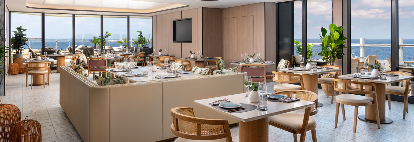 Bright, modern restaurant with wooden tables and chairs, neutral tones, and large windows offering ocean views. Tables are set for dining, some with patterned cushions. Green plants and natural light create a welcoming, airy atmosphere.