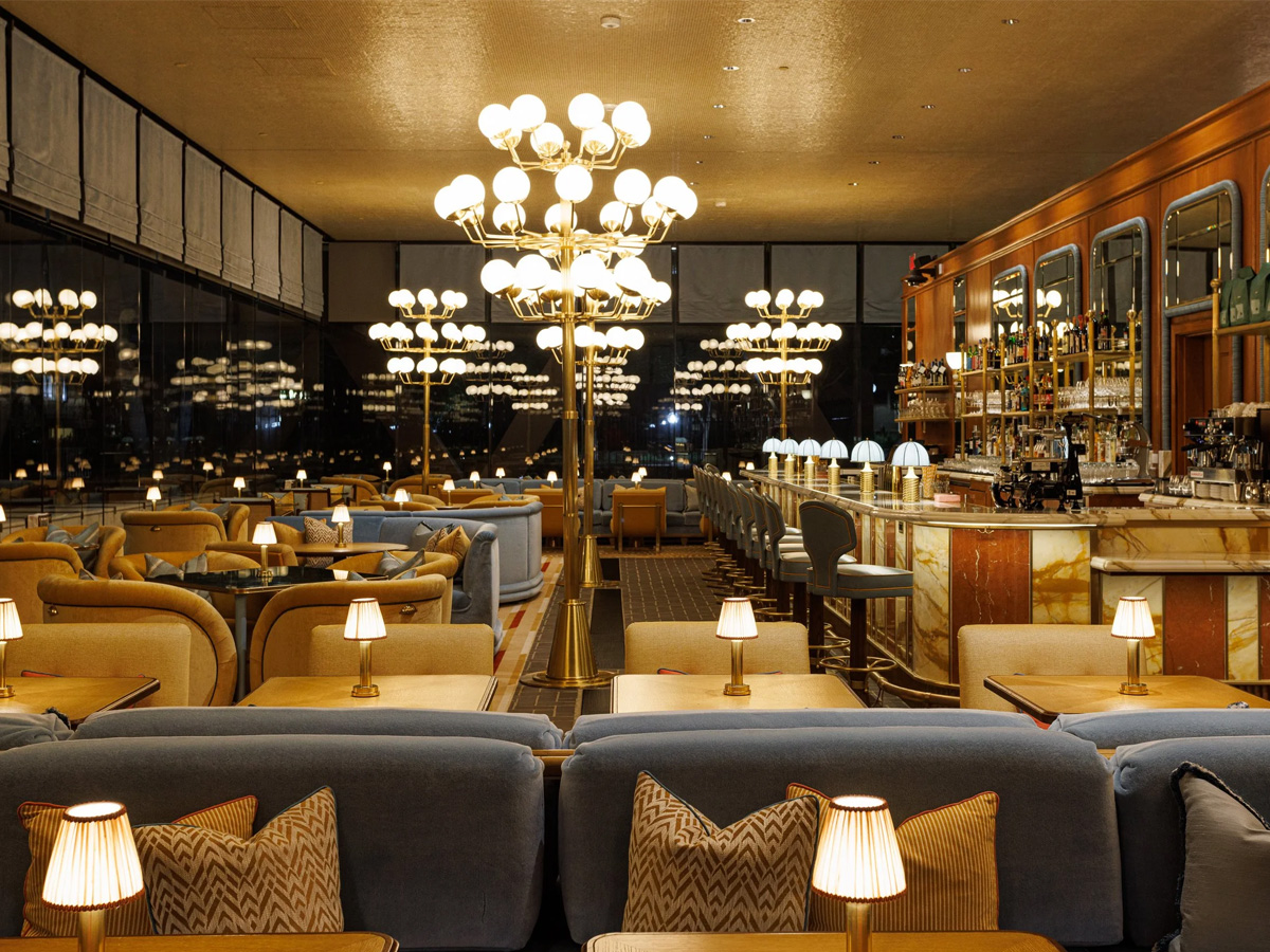 Elegant lounge with plush blue sofas, wooden tables, and small lamps. Ornate chandeliers hang from the ceiling. The bar, with marble accents and rows of bottles, is to the right. Large windows line the left side, revealing city lights outside at night.