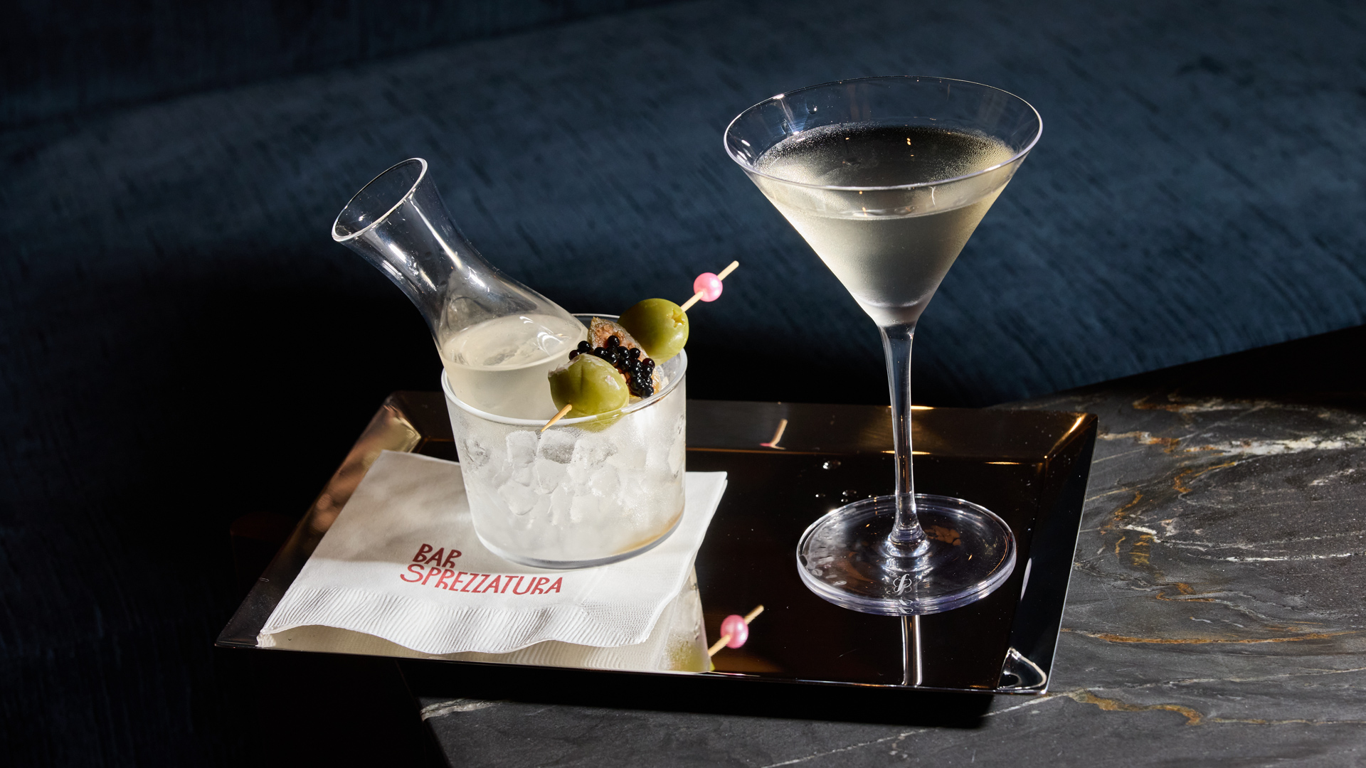 A martini glass and a small carafe with olives and caviar sit on a silver tray. The carafe is nestled in a glass bowl of ice atop a white napkin printed with Bar Sprezzatura. Both items rest on a dark, marble-patterned surface against a dark blue background.