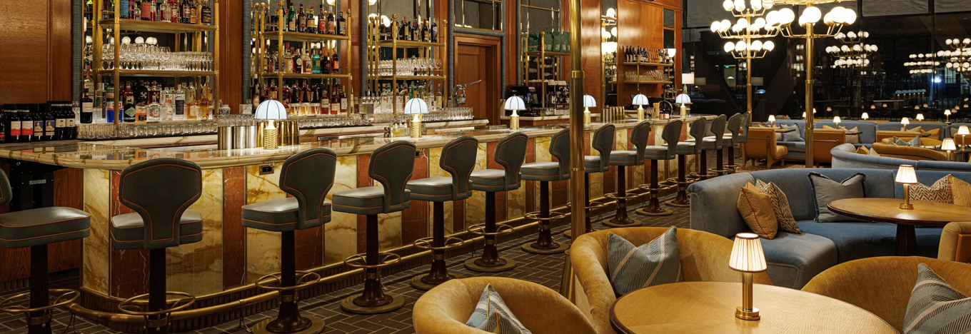 Elegant bar interior with a long marble counter lined with plush, high-backed barstools. Behind the bar are shelves of liquor and mirrors. Globe chandeliers, cozy booth seating, and round tables create a warm, inviting ambiance.