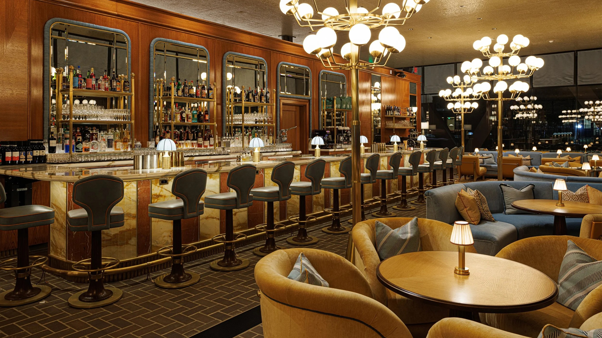 Elegant bar interior with a long marble counter lined with plush, high-backed barstools. Behind the bar are shelves of liquor and mirrors. Globe chandeliers, cozy booth seating, and round tables create a warm, inviting ambiance.