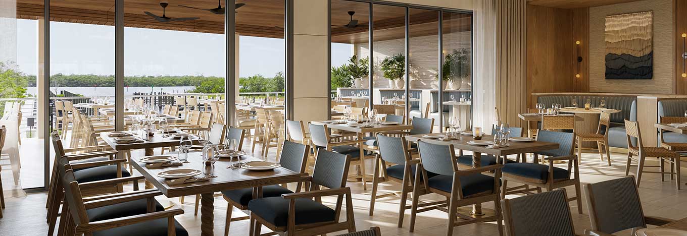Modern restaurant dining area with wooden tables and blue-cushioned chairs, set for service. Large windows and glass doors offer views of greenery and an outdoor patio with more seating. Light wood tones and natural light create an airy, inviting atmosphere.