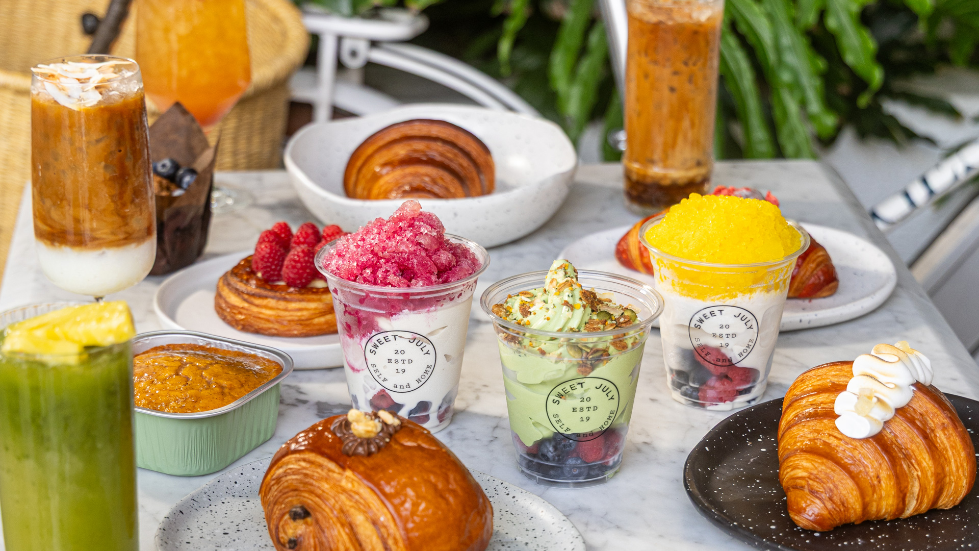 A marble table filled with assorted pastries and desserts, including croissants, pain au chocolat, fruit-topped shaved ice in cups, a muffin, and various iced drinks. Green plants and part of a wicker bag are visible in the background.