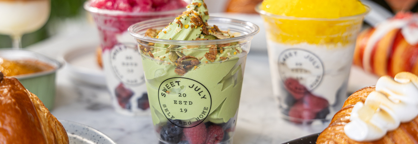 A plastic cup labeled “Sweet July” holds green matcha soft serve topped with granola and fruits, surrounded by similar dessert cups and pastries on a marble table.
