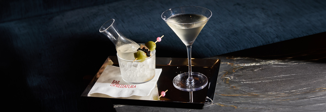 A martini and a small glass carafe with olives and caviar sit on a silver tray. The tray rests on a dark marble surface, and a napkin beneath the carafe is printed with the words Bar Sprezzatura in red.