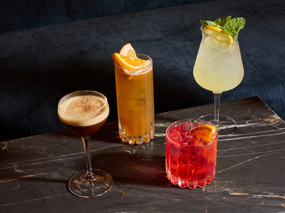 Four cocktails sit on a black marble table against a dark blue background. The drinks include a dark espresso-style martini, a tall orange-garnished amber cocktail, a light yellow drink with mint, and a bright red cocktail in a short glass with ice.