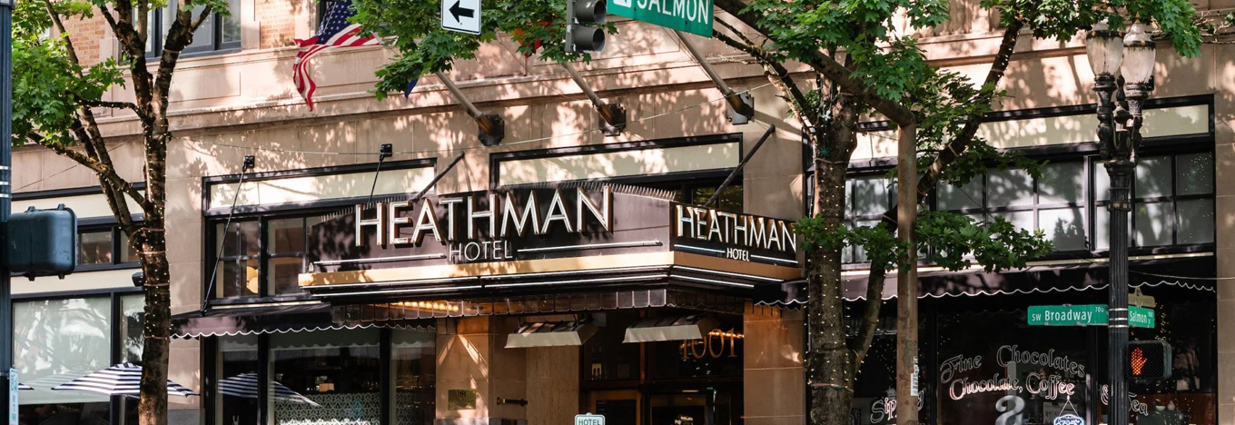 The Heathman Hotel entrance is shown behind leafy trees on a city street corner. The sign reads HEATHMAN HOTEL. An American flag hangs above the door. Two green street signs label the intersection as Broadway and Salmon.