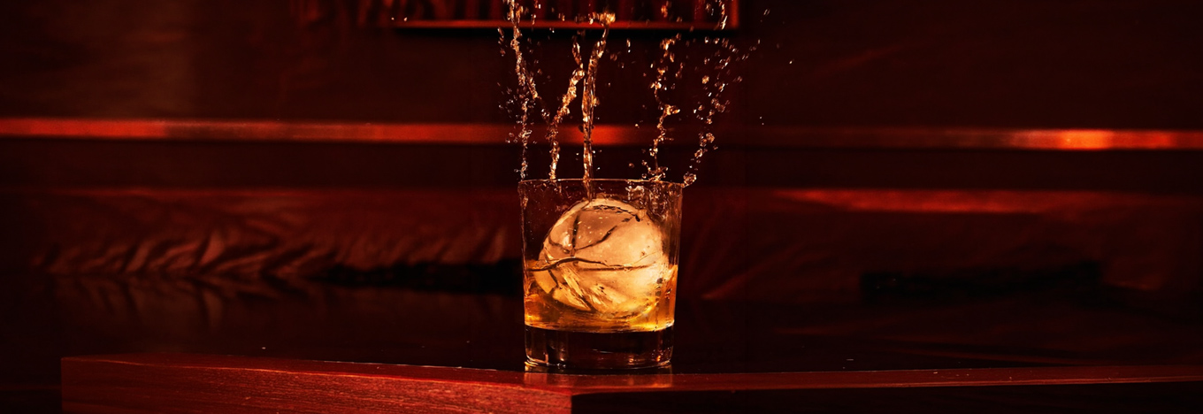 A short glass of whiskey with a large spherical ice cube sits on a dark wooden surface. Liquid splashes upward out of the glass in a dramatic spray, all set against a moody, dimly lit room with deep red tones.